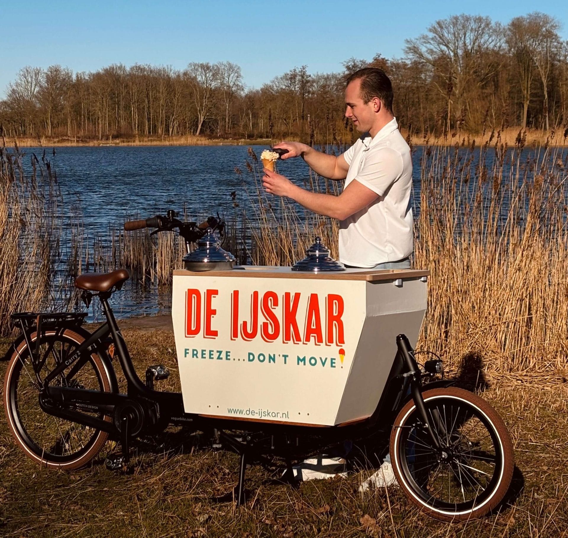 Ijscoman schept een hoorntje bij de ijskar-bakfiets aan de waterkant onder een strakblauwe lucht