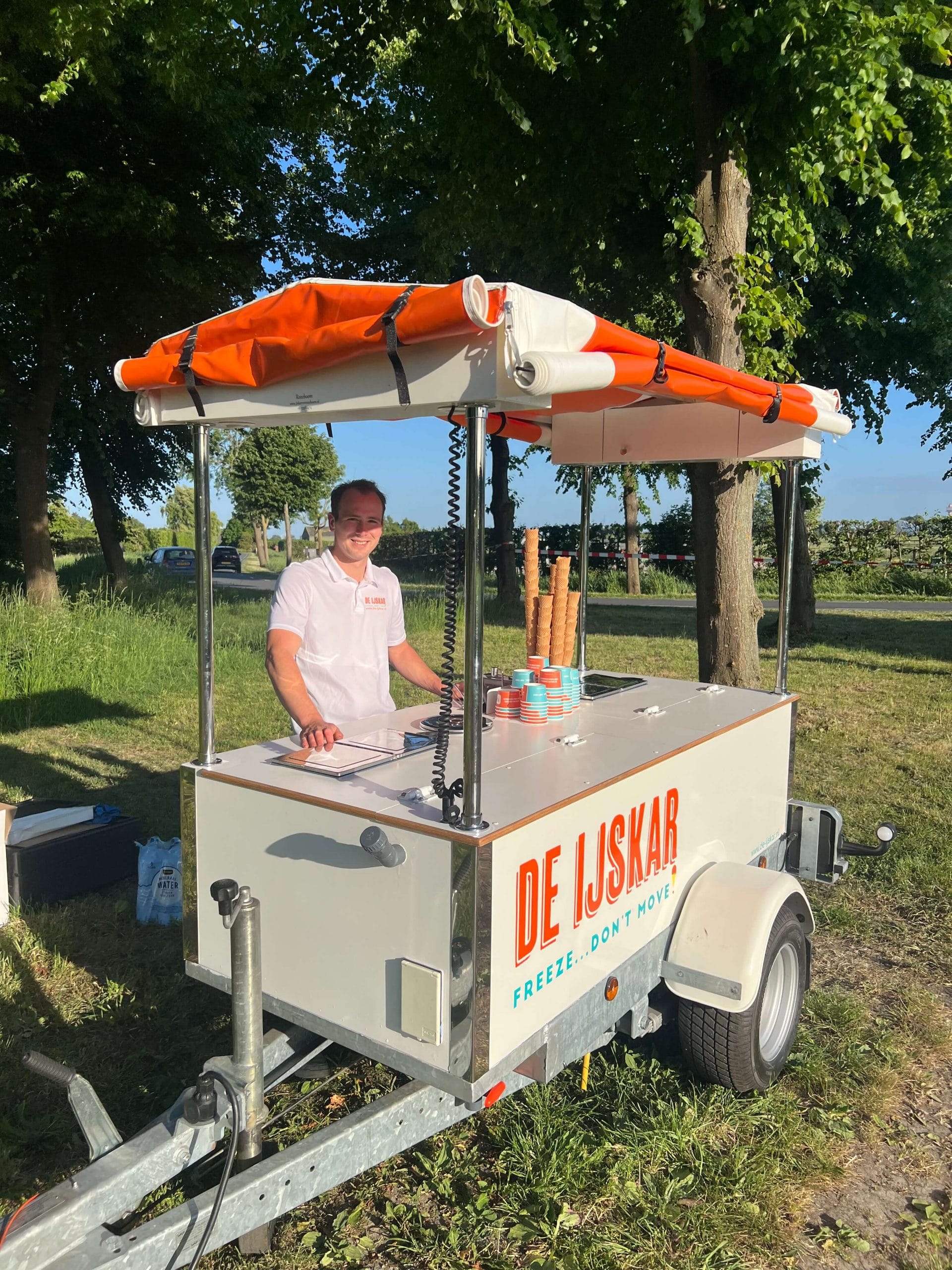De IJsfoodtruck met opgerolde oranje luifel op een grasveld, ijscoman achter de kar
